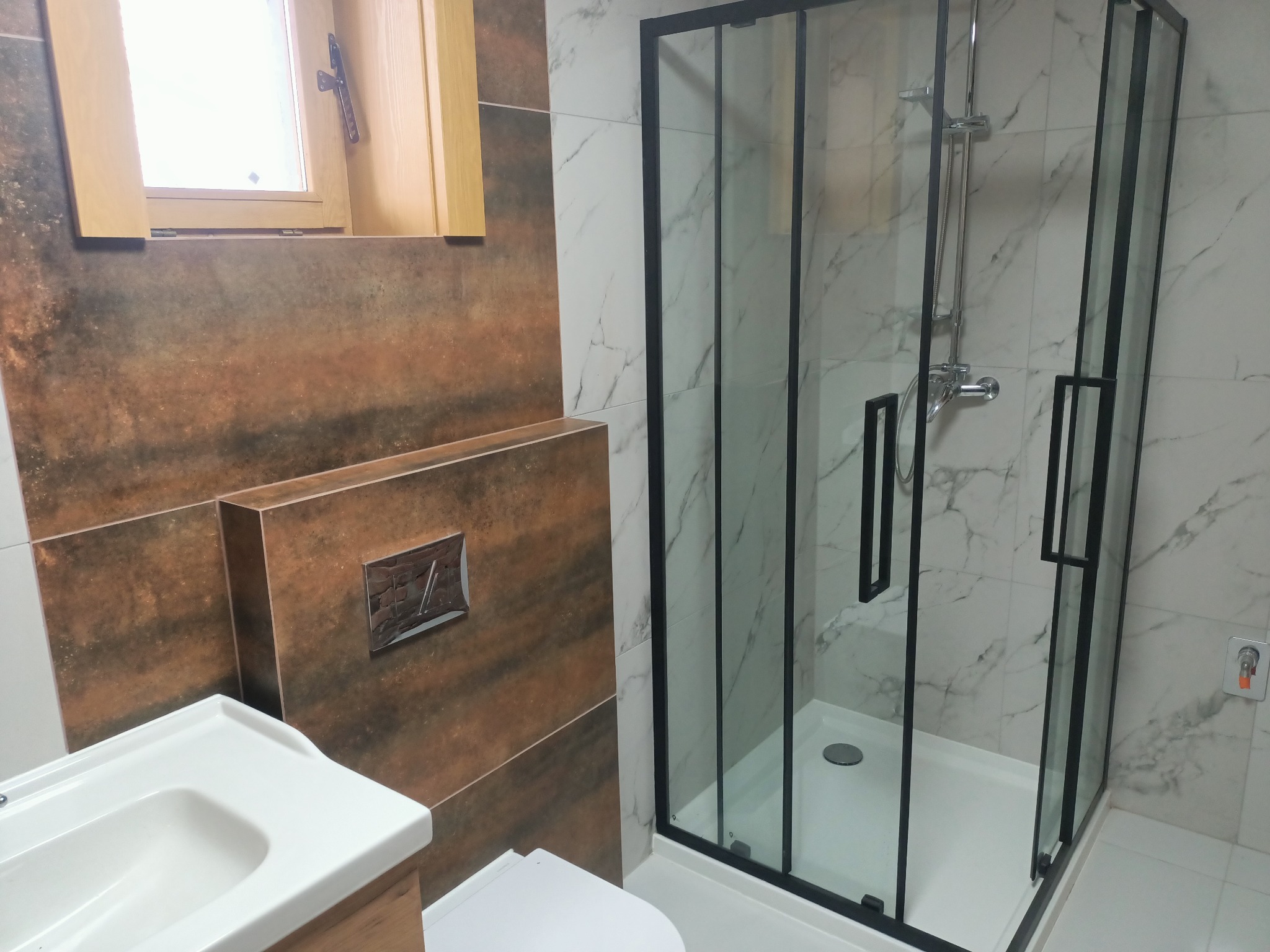 Nowoczesna łazienka z kabiną prysznicową w czarnej ramie, umywalką i toaletą. Ściany wyłożone płytkami imitującymi marmur i drewno. Okno z drewnianą ramą.