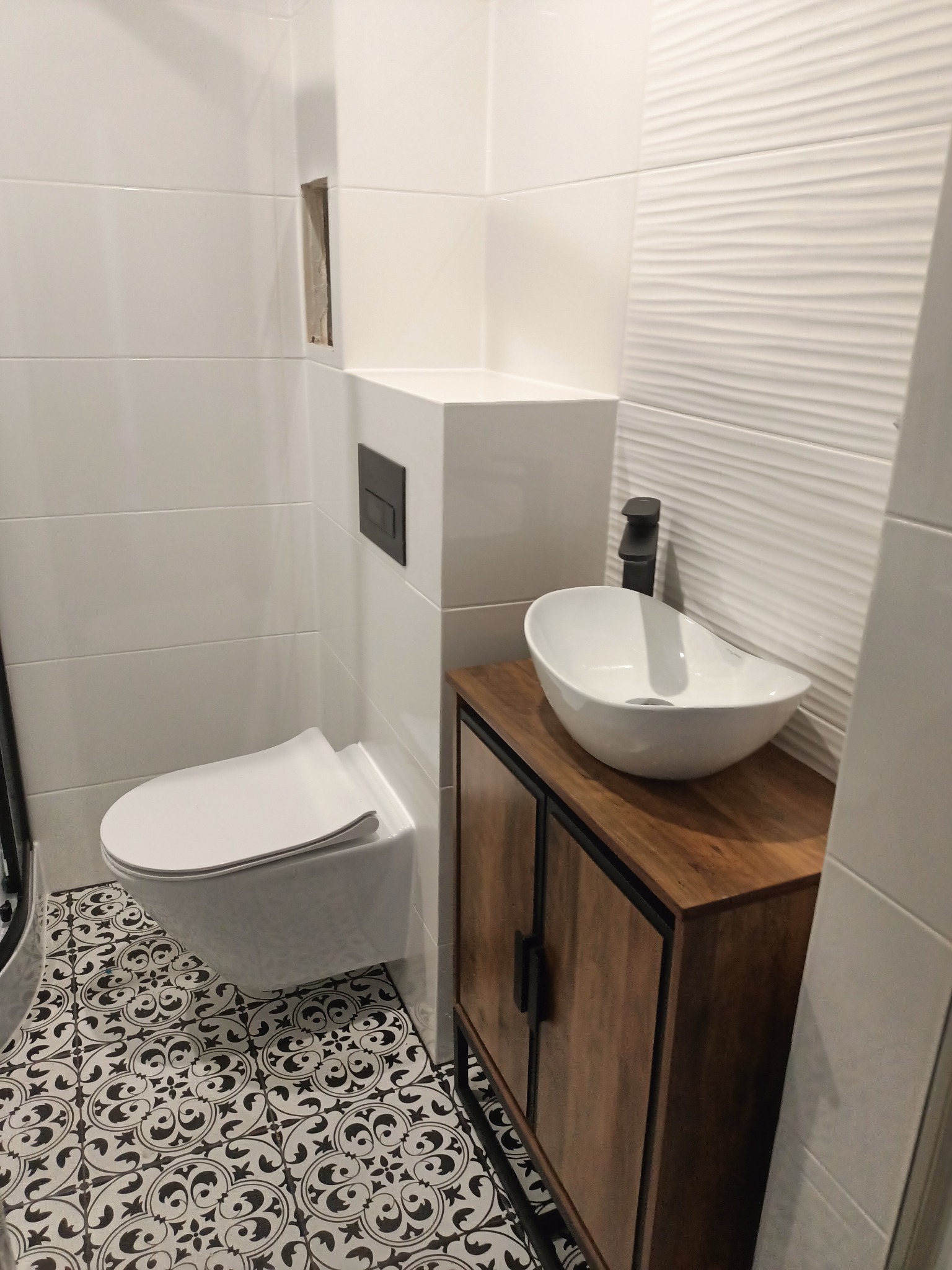 Nowoczesna łazienka z podwieszaną toaletą, umywalką nablatową na szafce z drewnianym frontem i czarno-białą wzorzystą podłogą. Białe płytki na ścianach, w tym ozdobne z fakturą.