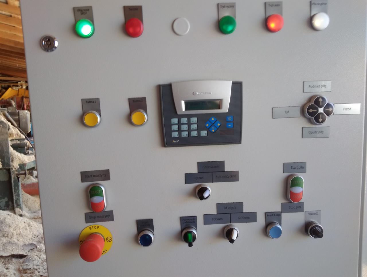 Szary panel sterowania maszyny z licznymi przyciskami, przełącznikami i wyświetlaczem, widoczne oznaczenia funkcji w języku polskim, w tle resztki drewna.
