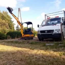 Żółta mini koparka JCB z podniesioną łyżką obok białego samochodu dostawczego Volkswagen na trawiastym terenie.