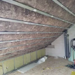 Poddasze w trakcie izolacji wełną mineralną, widoczne metalowe profile konstrukcyjne i płyty gipsowo-kartonowe przygotowane do montażu.