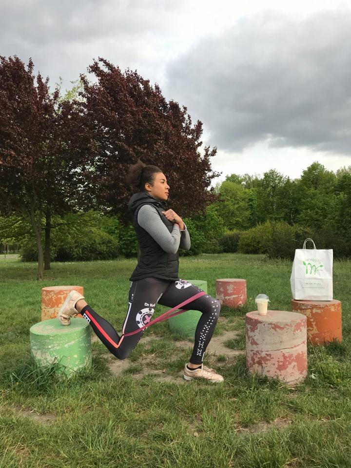 Kobieta wykonuje wypad w przód z gumą oporową na tle parku, obok stoi smoothie i torba na jedzenie.