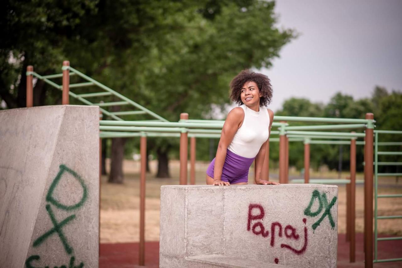 Uśmiechnięta kobieta w sportowym stroju opiera się o betonowy element placu do street workoutu, w tle drabinki i drzewa.