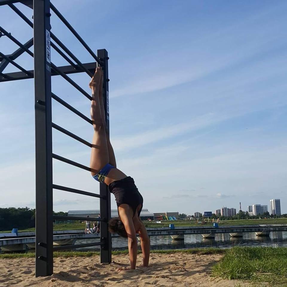 Kobieta w pozycji odwróconej, stojąca na rękach przy drabinkach na plaży, w tle jezioro i budynki miejskie.