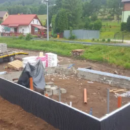 Fundamenty domu w trakcie budowy, zabezpieczone czarną folią izolacyjną, widoczny transport kostki brukowej na przyczepie za białym dostawczym samochodem, w tle zabudowa jednorodzinna i zieleń.