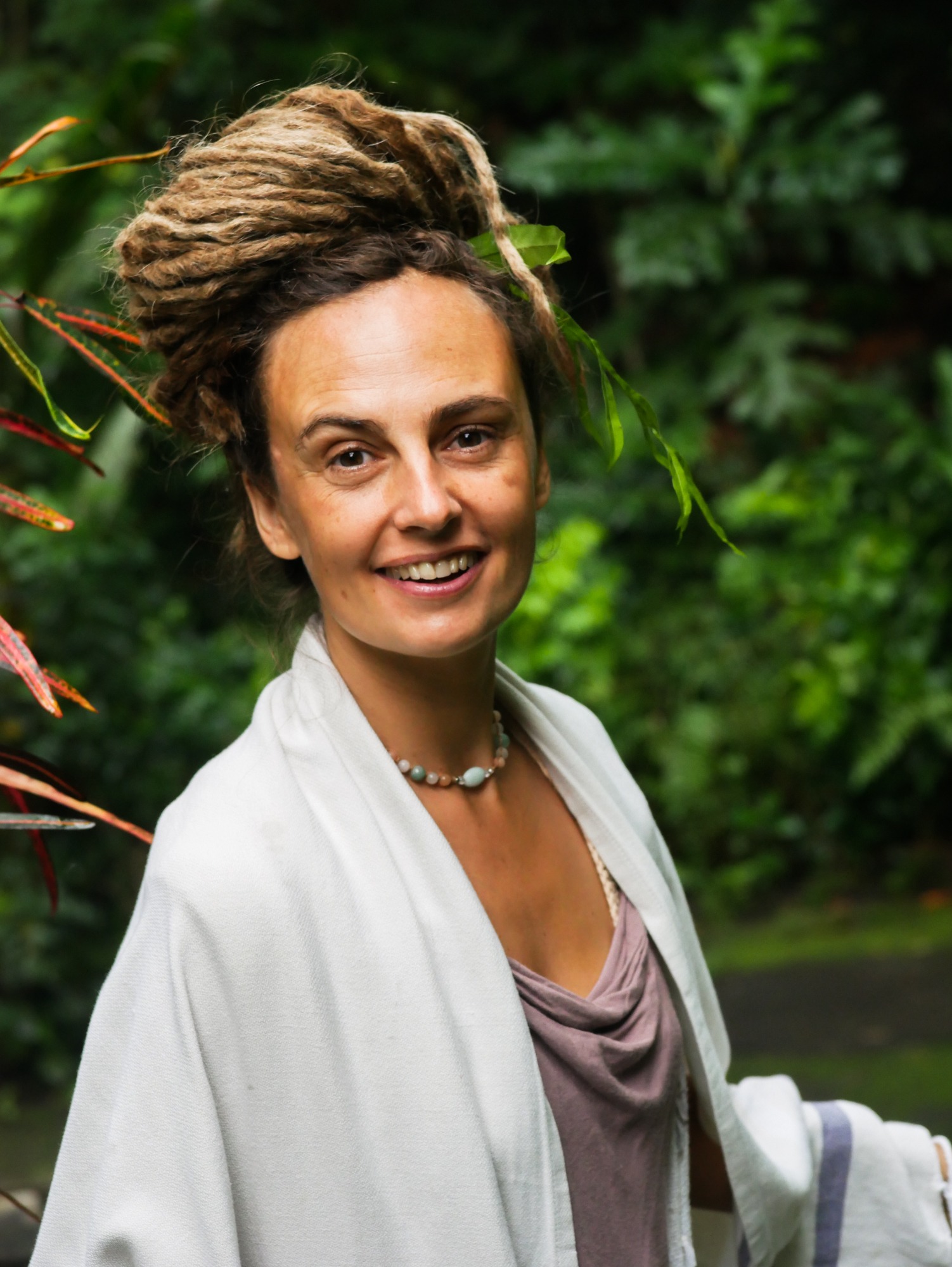 Portret uśmiechniętej kobiety z dredami upiętymi na czubku głowy, z liśćmi wplecionymi we włosy, ubrana w jasny szal i naszyjnik z kamieni, na tle bujnej zieleni.
