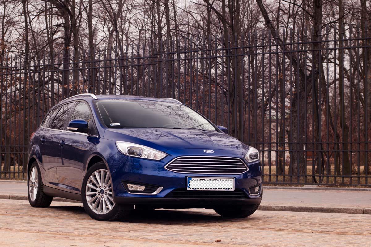 Niebieski Ford Focus kombi zaparkowany na brukowanej ulicy z metalowym ogrodzeniem i drzewami bez liści w tle.