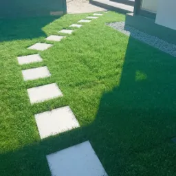 Ścieżka z jasnych, kwadratowych płyt ułożona w trawniku, obok rabata wysypana drobnymi kamykami, widok z góry.