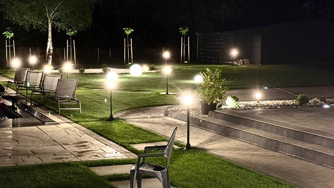 Oświetlony ogród nocą: ścieżka z kostki, trawnik, lampy kuliste, drzewa i meble ogrodowe tworzą magiczną atmosferę. Widoczne schody i rośliny w donicach.
