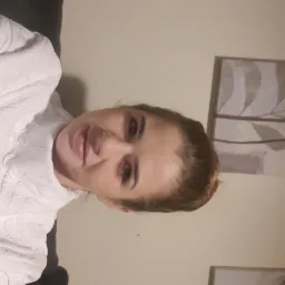 Portret kobiety w białym swetrze z abstrakcyjnym obrazem roślinnym w tle, uchwycony w słabym oświetleniu.