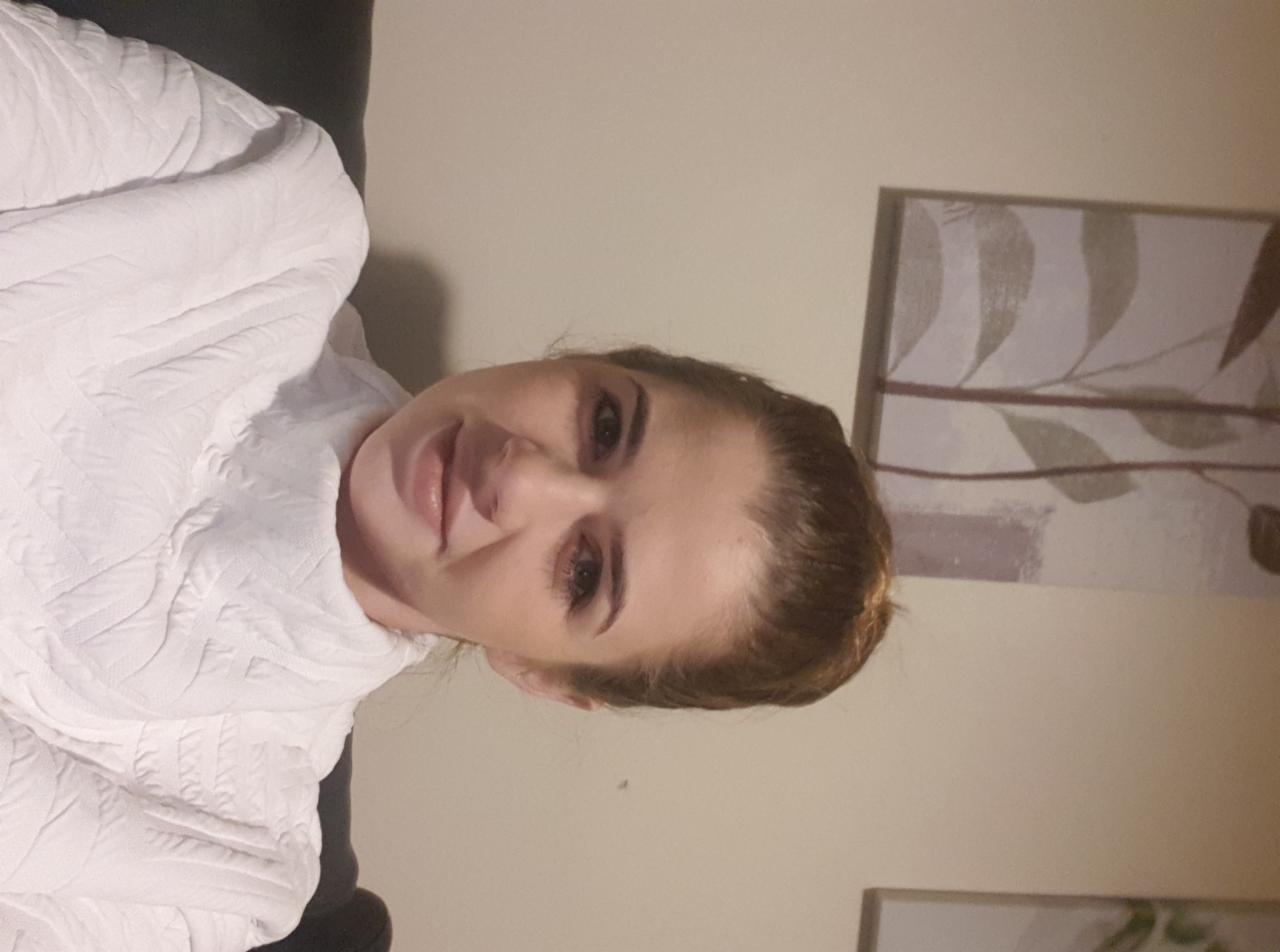 Portret kobiety w białym swetrze z abstrakcyjnym obrazem roślinnym w tle, uchwycony w słabym oświetleniu.