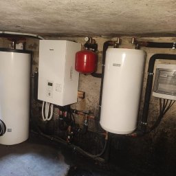 STROMER energia słoneczna i pompy ciepła - Biały, wiszący piec gazowy Panasonic, dwa cylindryczne zbiorniki na wodę i czerwony zbiornik wyrównawczy w kotłowni z instalacją, rurami i skrzynką elektryczną.