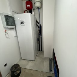 STROMER energia słoneczna i pompy ciepła - Biała pompa ciepła LG Therma V w pomieszczeniu technicznym, z widoczną instalacją hydrauliczną i elektryczną, oraz zbiornikami wyrównawczymi. Surowy stan wykończenia.