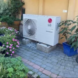 Biała pompa ciepła LG Therma V, ustawiona na betonowych blokach w ogrodzie, otoczona kwiatami i roślinami w donicach. Widoczny fragment roweru.