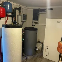 W kotłowni biały zbiornik z czerwonym i niebieskim naczyniem wzbiorczym, szary zbiornik oraz biały panel sterowania Fox. Widoczna instalacja rurowa i skrzynki elektryczne.