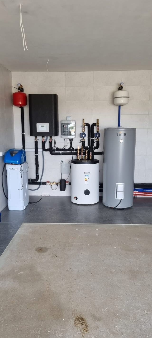 Kompletna instalacja grzewcza z dwoma zbiornikami, kotłem i filtrem wody w pomieszczeniu wyłożonym płytkami. Widoczne rury i zawory.