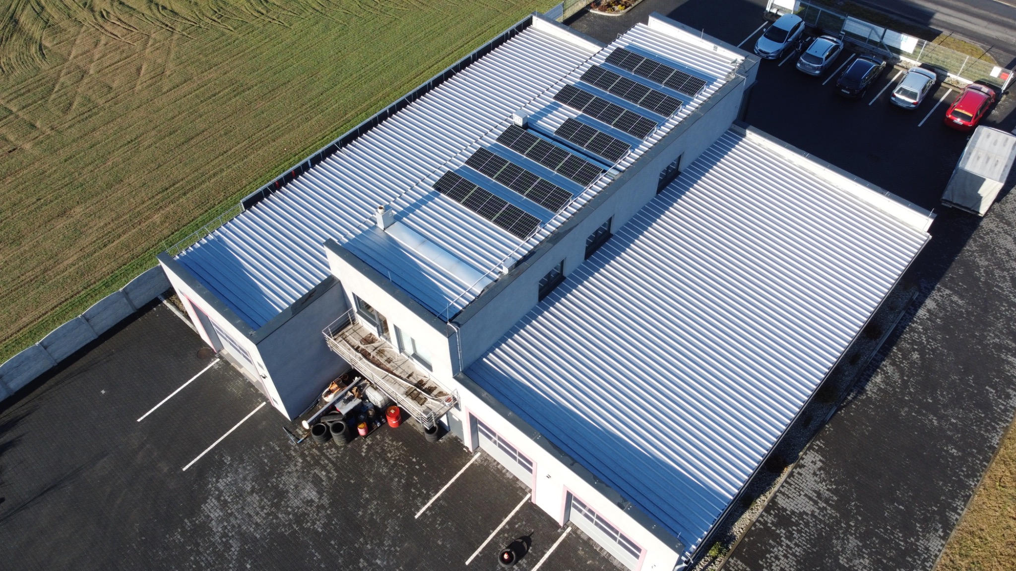 Widok z drona na budynek z panelami słonecznymi na dachu, obok pole uprawne i parking z samochodami. Panele ułożone w rzędach, dach blaszany trapezowy.