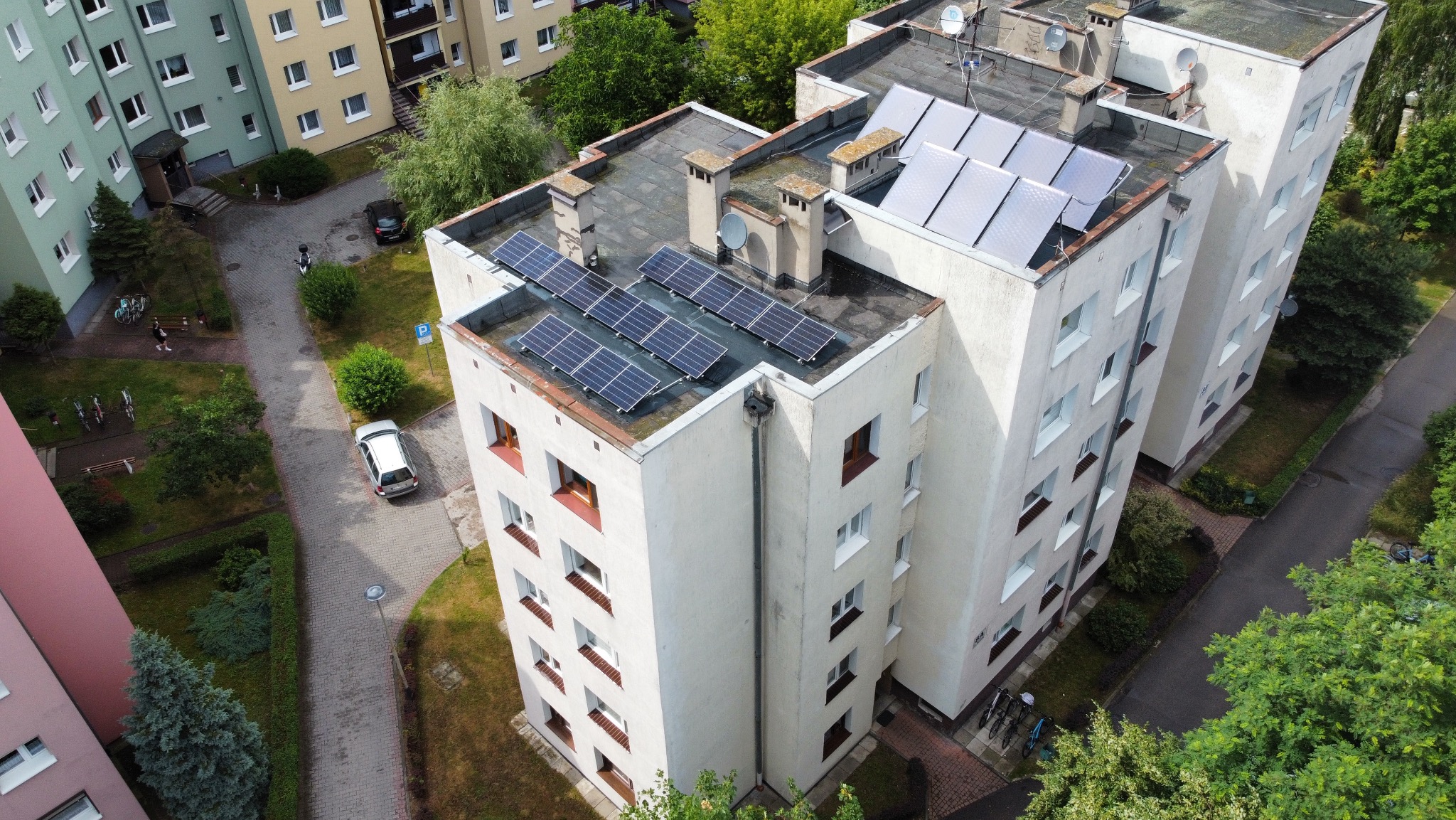 Widok z góry na budynek wielorodzinny z panelami słonecznymi na dachu. Panele ułożone w dwóch sekcjach, widoczne kominy wentylacyjne, drzewa i zaparkowane samochody.