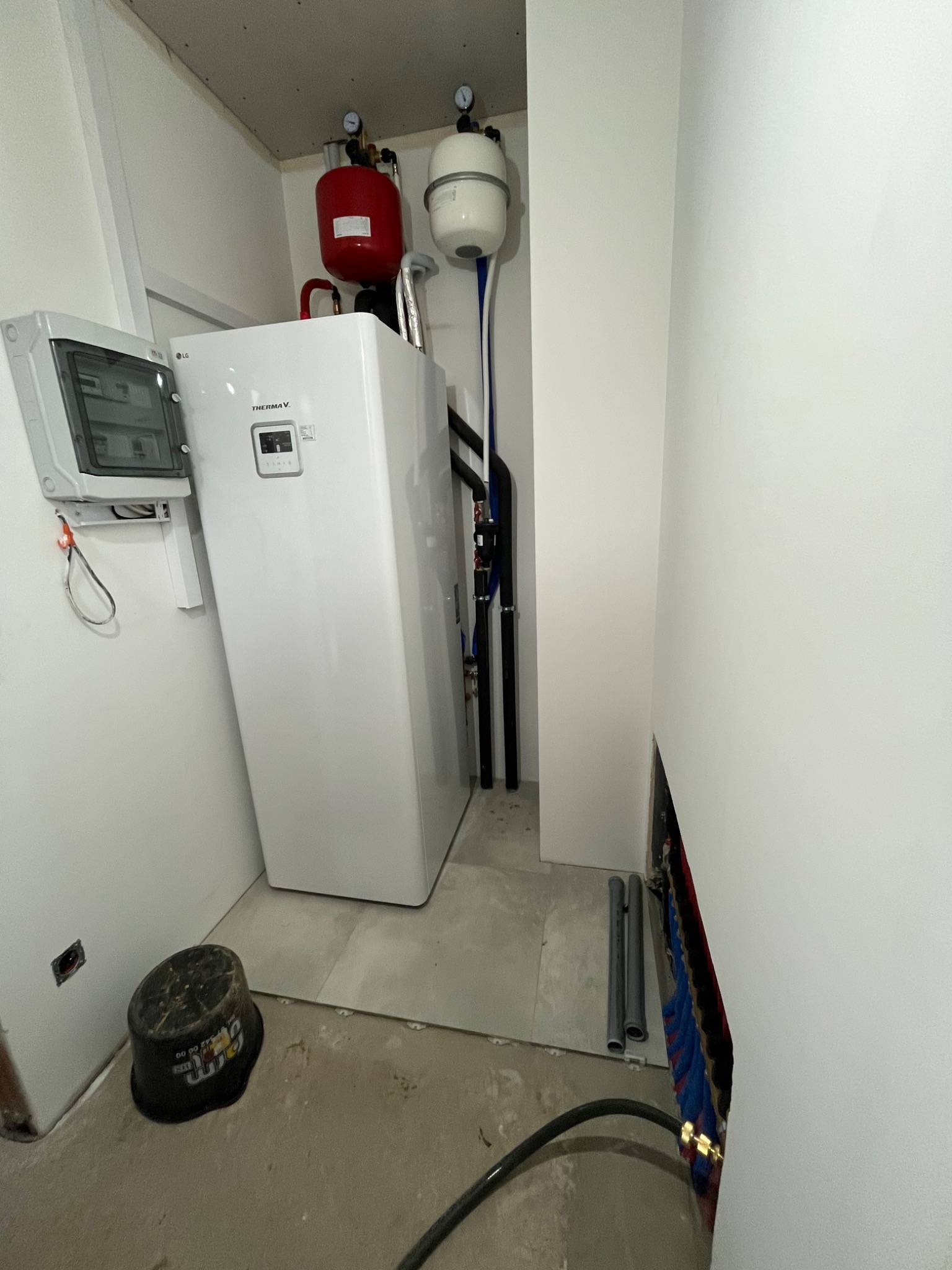Biała pompa ciepła LG Therma V w pomieszczeniu technicznym, z widoczną instalacją hydrauliczną i elektryczną, oraz zbiornikami wyrównawczymi. Surowy stan wykończenia.