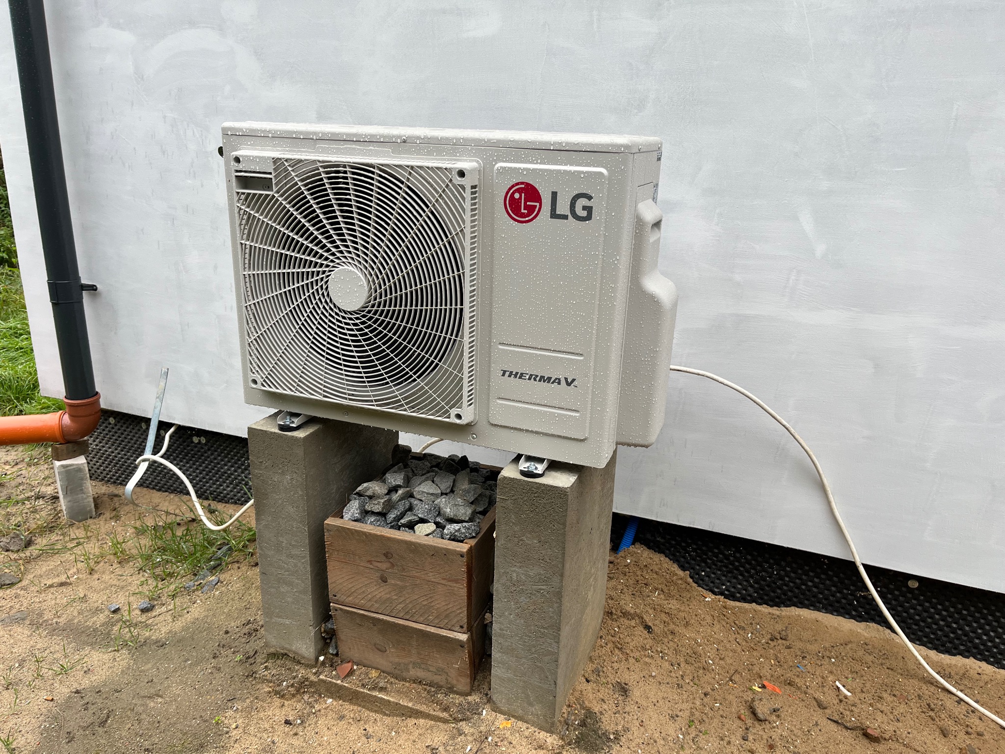 Zamontowana jednostka zewnętrzna pompy ciepła LG Therma V na betonowych wspornikach, z widoczną instalacją elektryczną i elementami otoczenia budynku w deszczowy dzień.