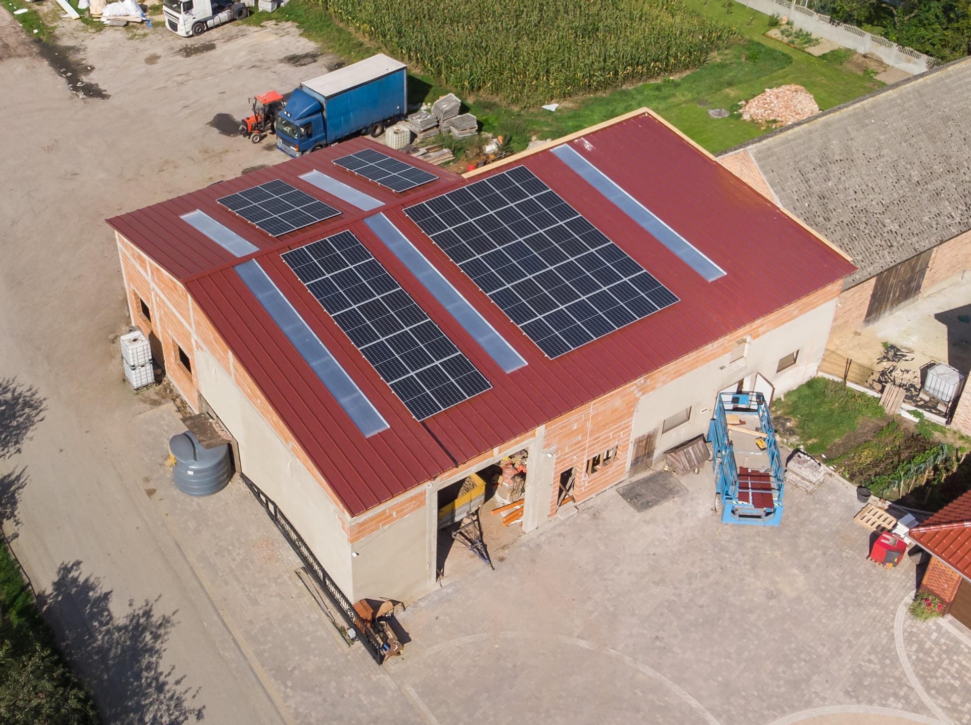Widok z góry na budynek z panelami słonecznymi na czerwonym dachu, obok pole kukurydzy, częściowo otynkowany, z oknami dachowymi i maszynami budowlanymi obok.