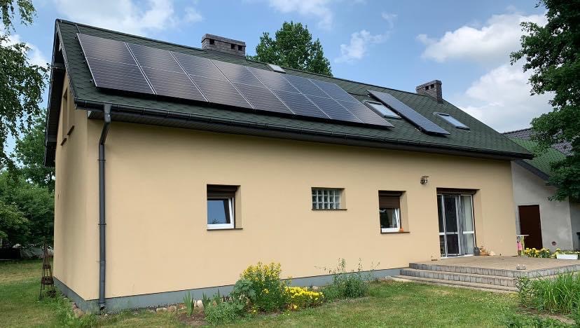 Dom jednorodzinny z panelami słonecznymi na zielonym dachu, widok z boku, jasne niebo, ogród z kwitnącymi żółtymi kwiatami.