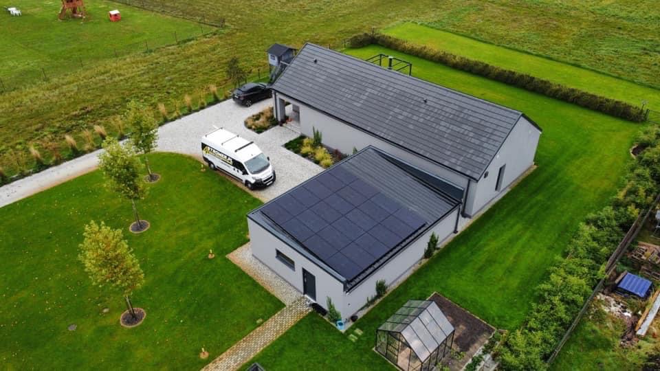 Nowoczesny dom z panelami słonecznymi na dachu, widok z góry. Na podjeździe stoi biały bus firmy instalacyjnej. Zadbany trawnik i szklarnia w ogrodzie.