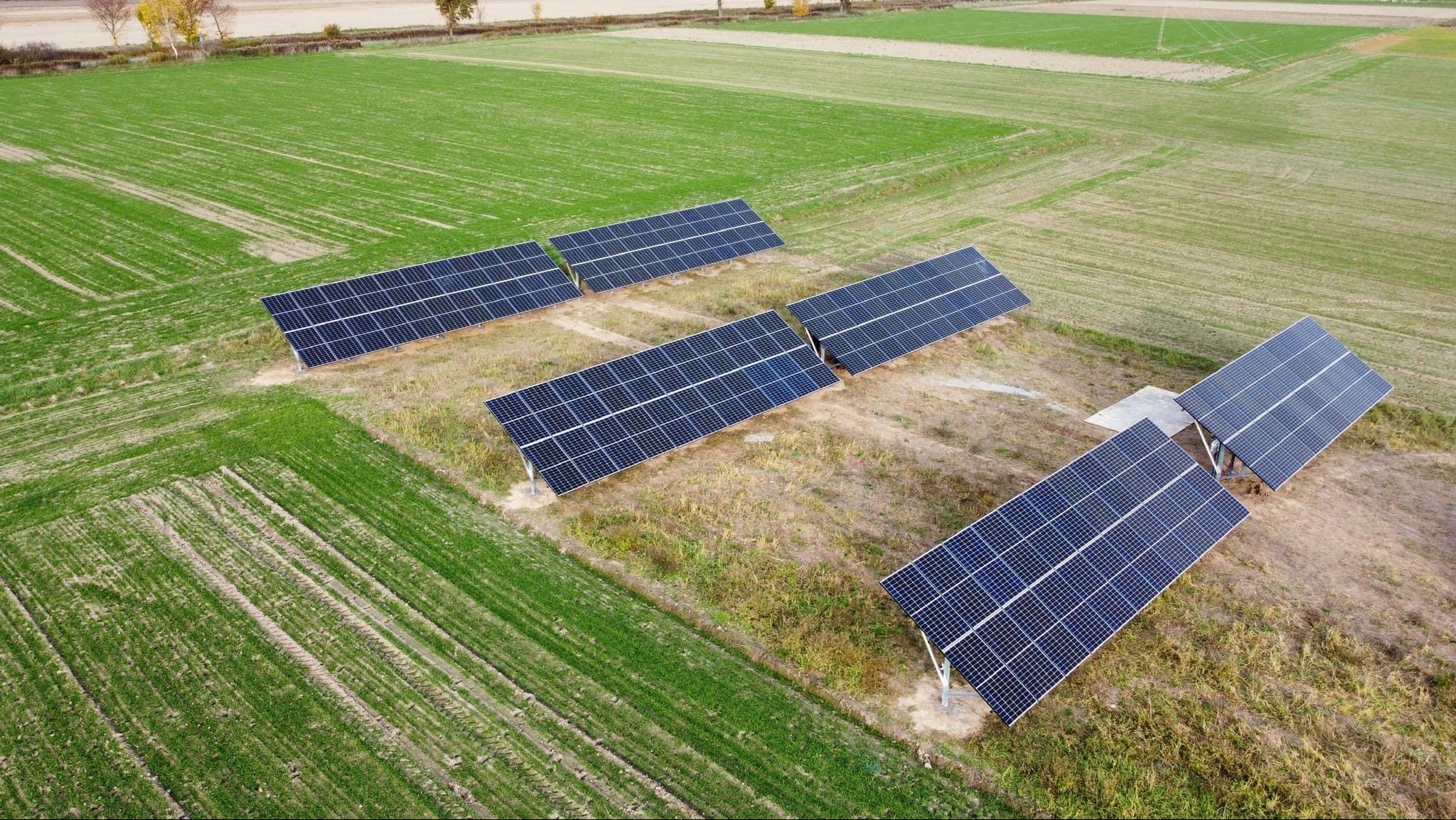Widok z góry na instalację fotowoltaiczną na polu uprawnym. Panele w rzędach, z wyraźnymi śladami po przejazdach maszyn rolniczych, betonowa podstawa pod jednym z paneli.