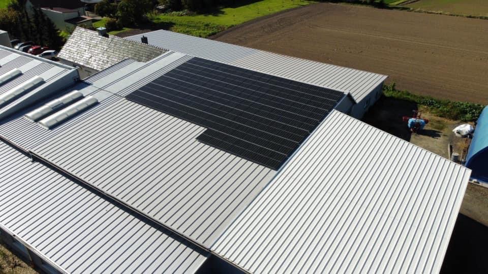 Widok z góry na dach budynku gospodarczego z zamontowanymi panelami fotowoltaicznymi. Dach blaszany, trapezowy, w kolorze srebrnym. W tle pola uprawne i zabudowa.