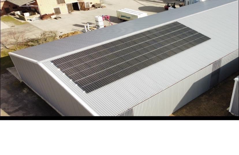 Panele słoneczne na dachu magazynu z blachy falistej, widok z góry. Na drugim planie budynek gospodarczy i plac manewrowy z pojazdami.