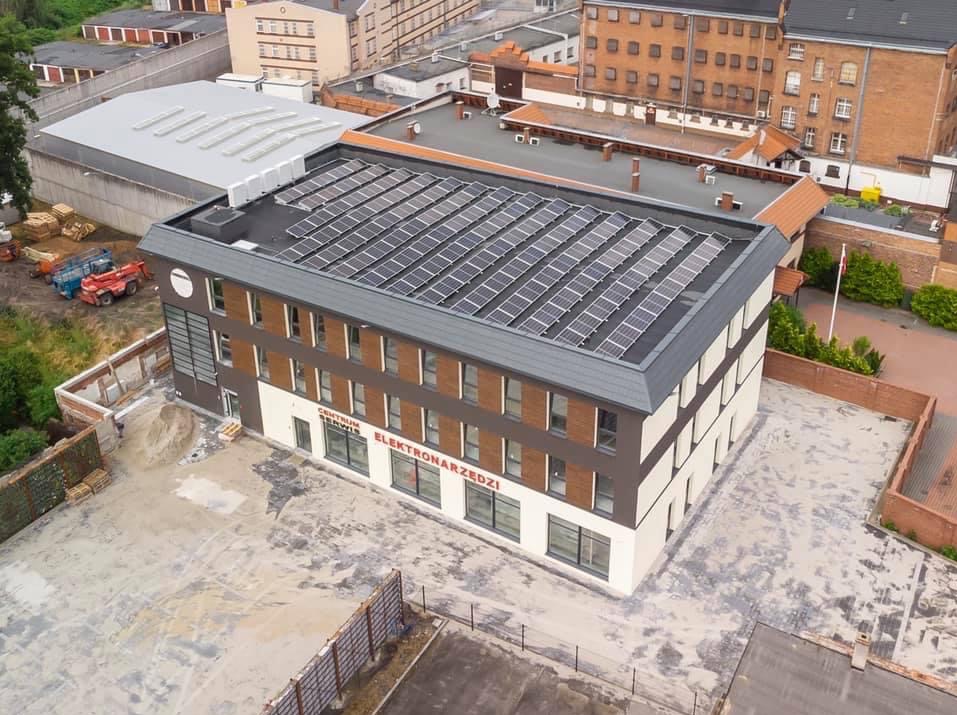 Widok z góry na budynek z panelami słonecznymi na dachu, plac budowy obok i budynki w tle. Centrum Serwis Elektronarzędzi.
