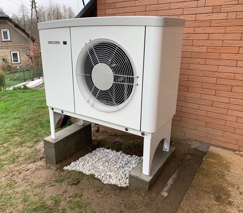 Biała pompa ciepła marki Stiebel Eltron, zamontowana na betonowych fundamentach obok ceglanego budynku. Pod urządzeniem wysypany żwir.