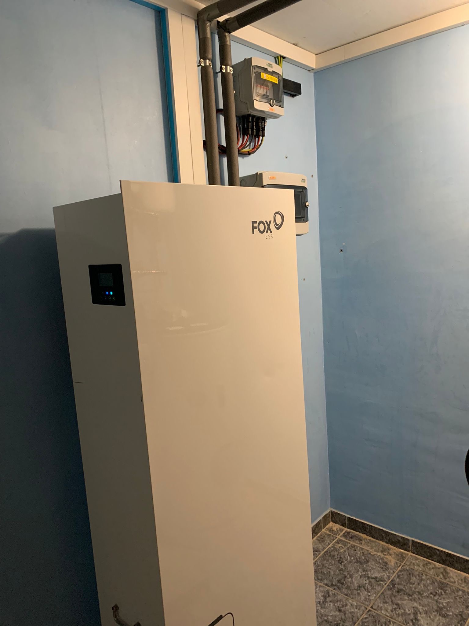 Biała jednostka Fox ESS w pomieszczeniu z rurami i skrzynkami elektrycznymi na niebieskiej ścianie. Widoczny fragment podłogi z szarej terakoty.
