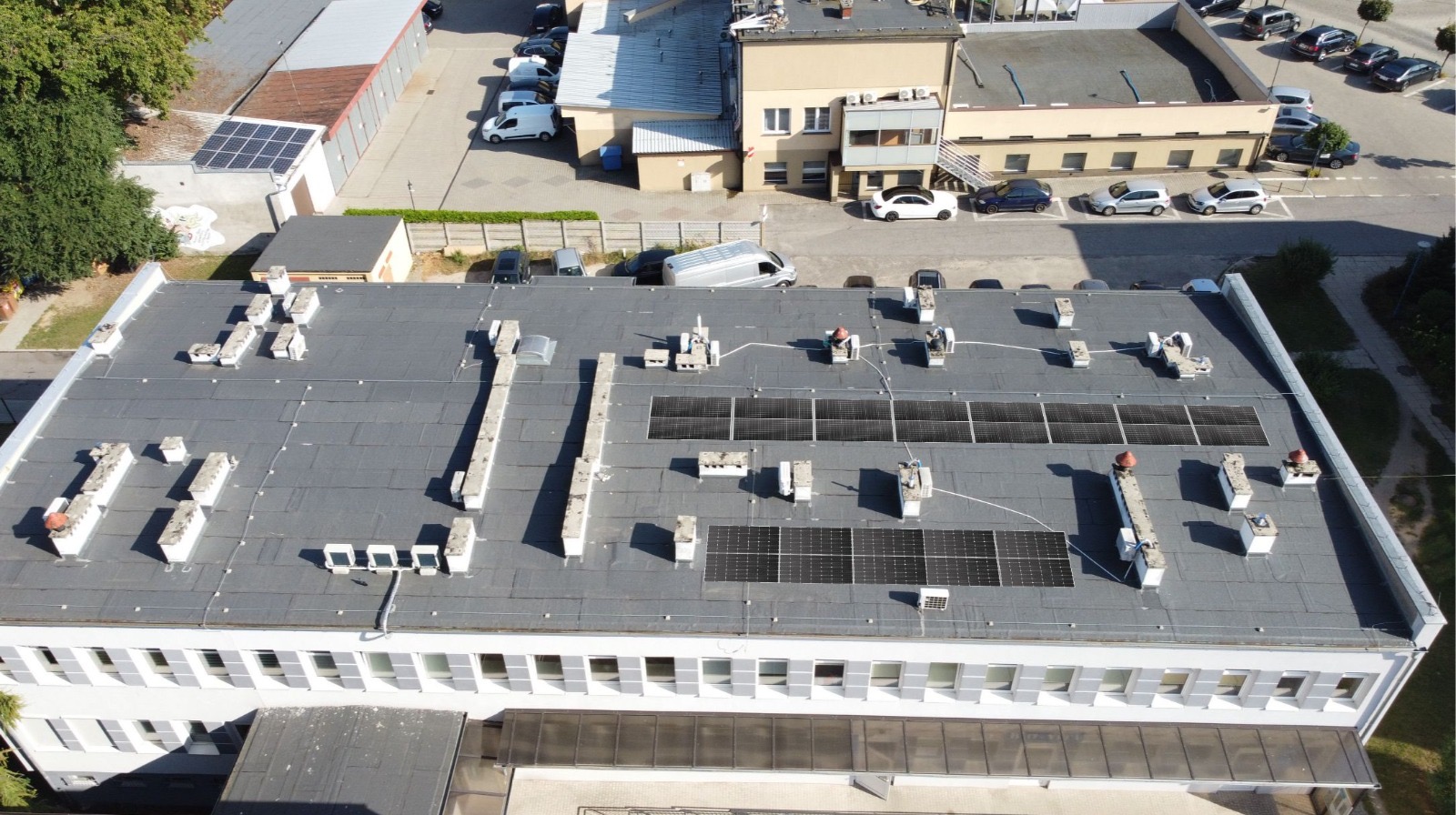 Widok z góry na płaski dach budynku z zainstalowanymi panelami fotowoltaicznymi i elementami wentylacji. Na dachu widoczne również świetliki dachowe.