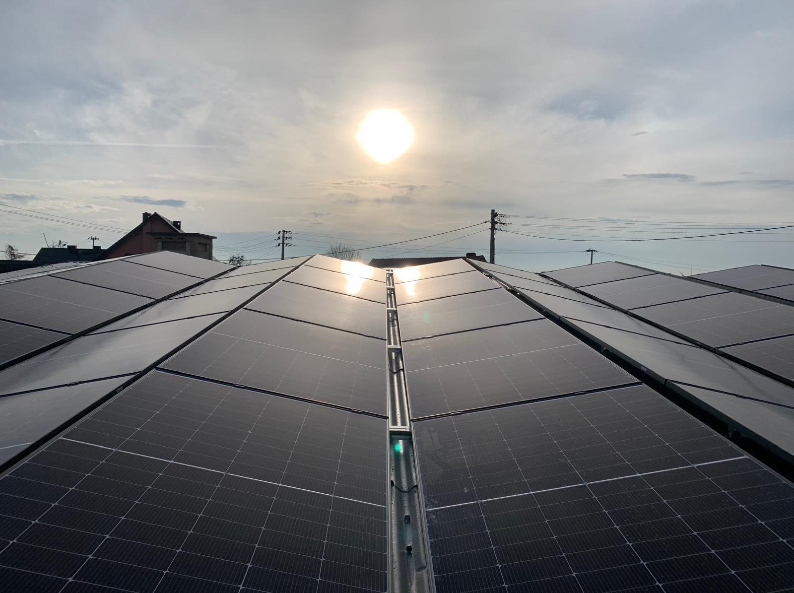 Błyszczące panele fotowoltaiczne na dachu w promieniach słońca, widok z góry. W tle budynek i słupy energetyczne.