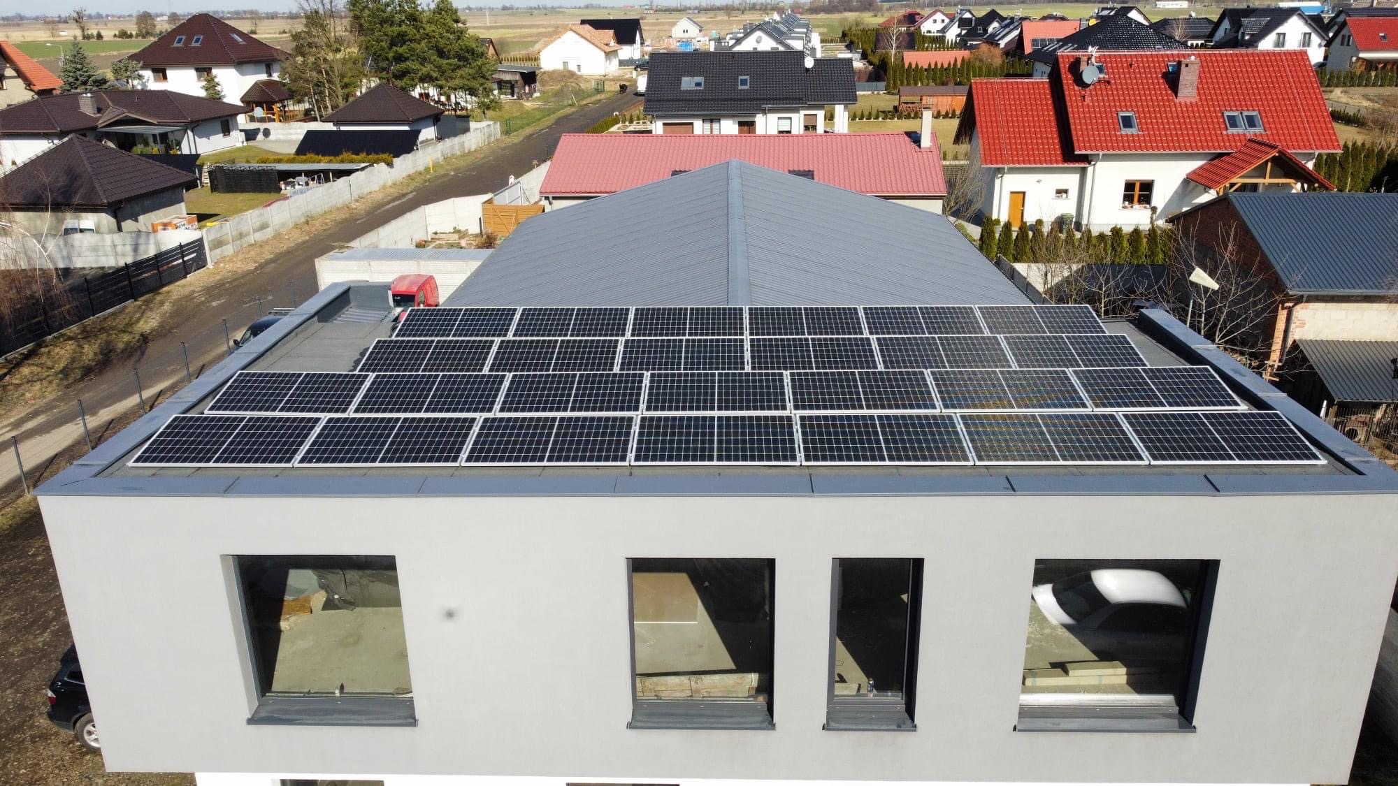 Panele słoneczne na dachu nowoczesnego, dwukondygnacyjnego domu z płaskim dachem. Widok z góry na instalację fotowoltaiczną w otoczeniu zabudowy jednorodzinnej.