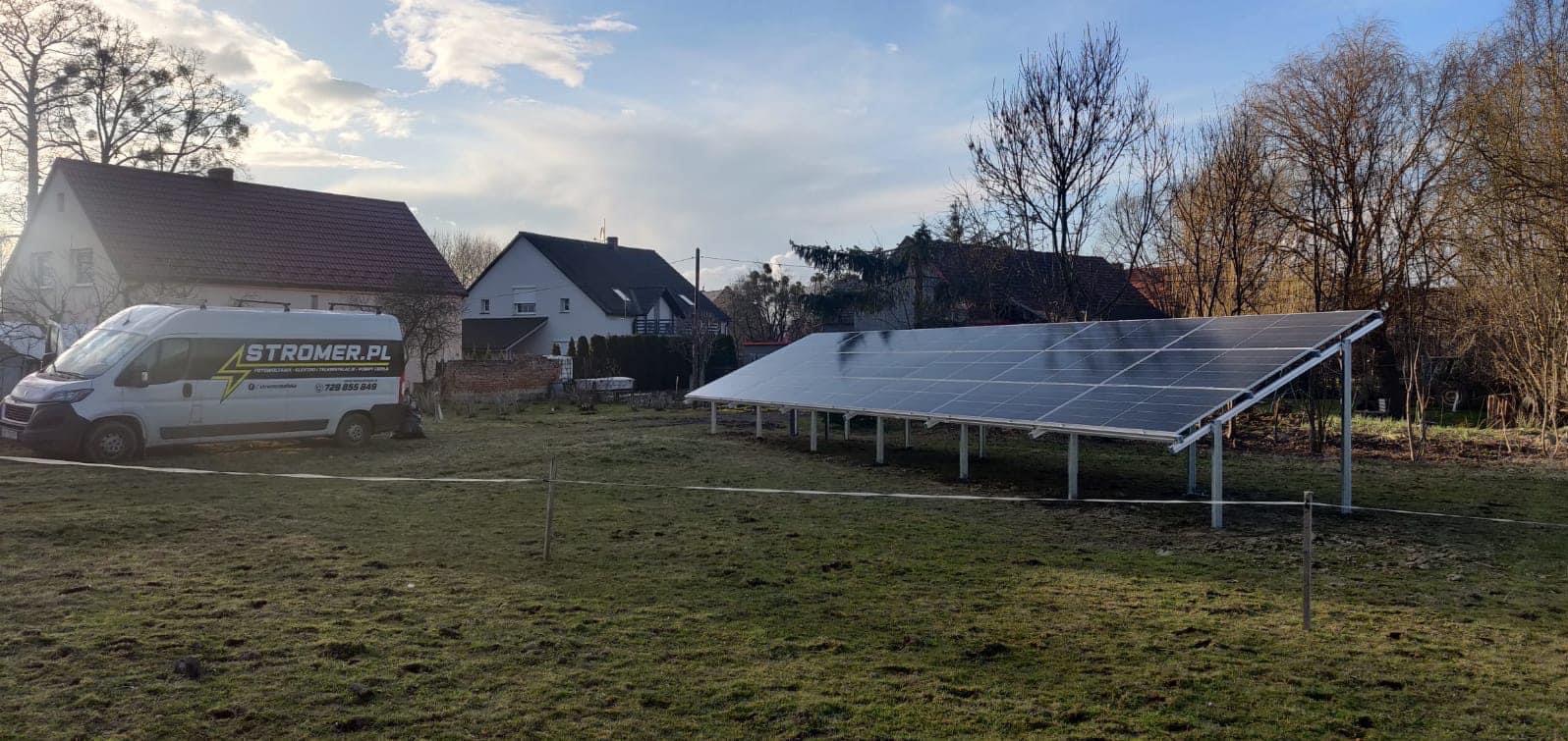 Instalacja fotowoltaiczna na gruncie, panele ustawione na metalowych wspornikach. W tle budynki mieszkalne i bus firmy Stromer.pl.