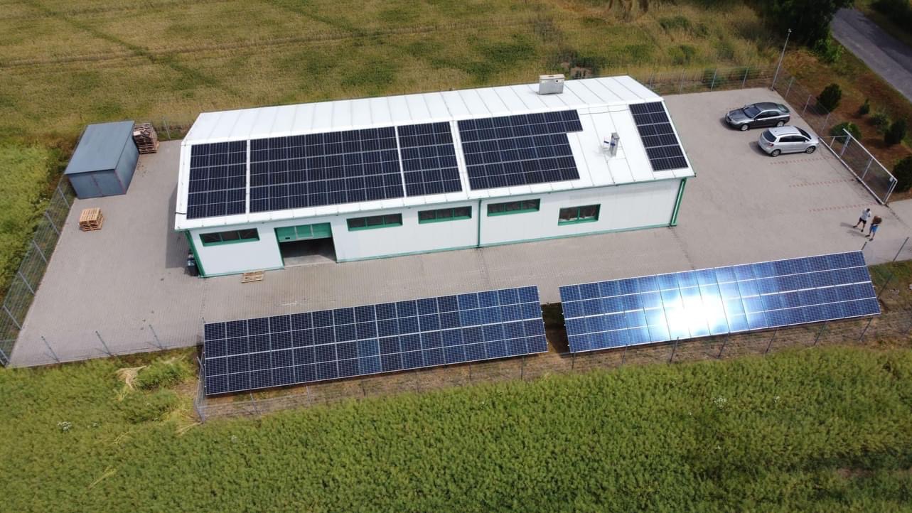 Widok z góry na budynek z panelami słonecznymi na dachu i na gruncie. Dwa samochody zaparkowane obok. Obok budynku widoczna zieleń.