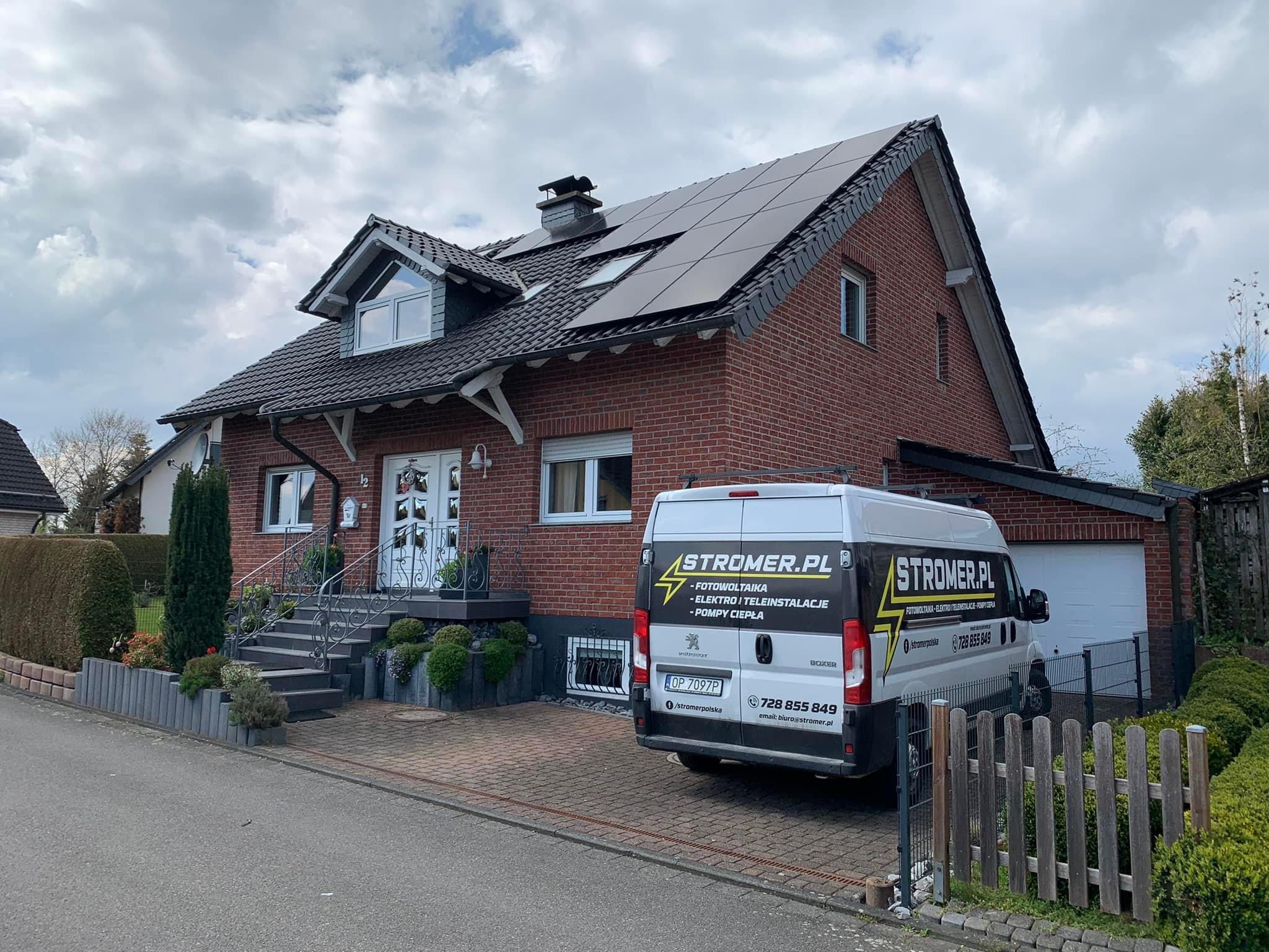 Dom z panelami słonecznymi na dachu, na podjeździe zaparkowany bus firmy instalującej fotowoltaikę. Widok na fasadę budynku i otoczenie.