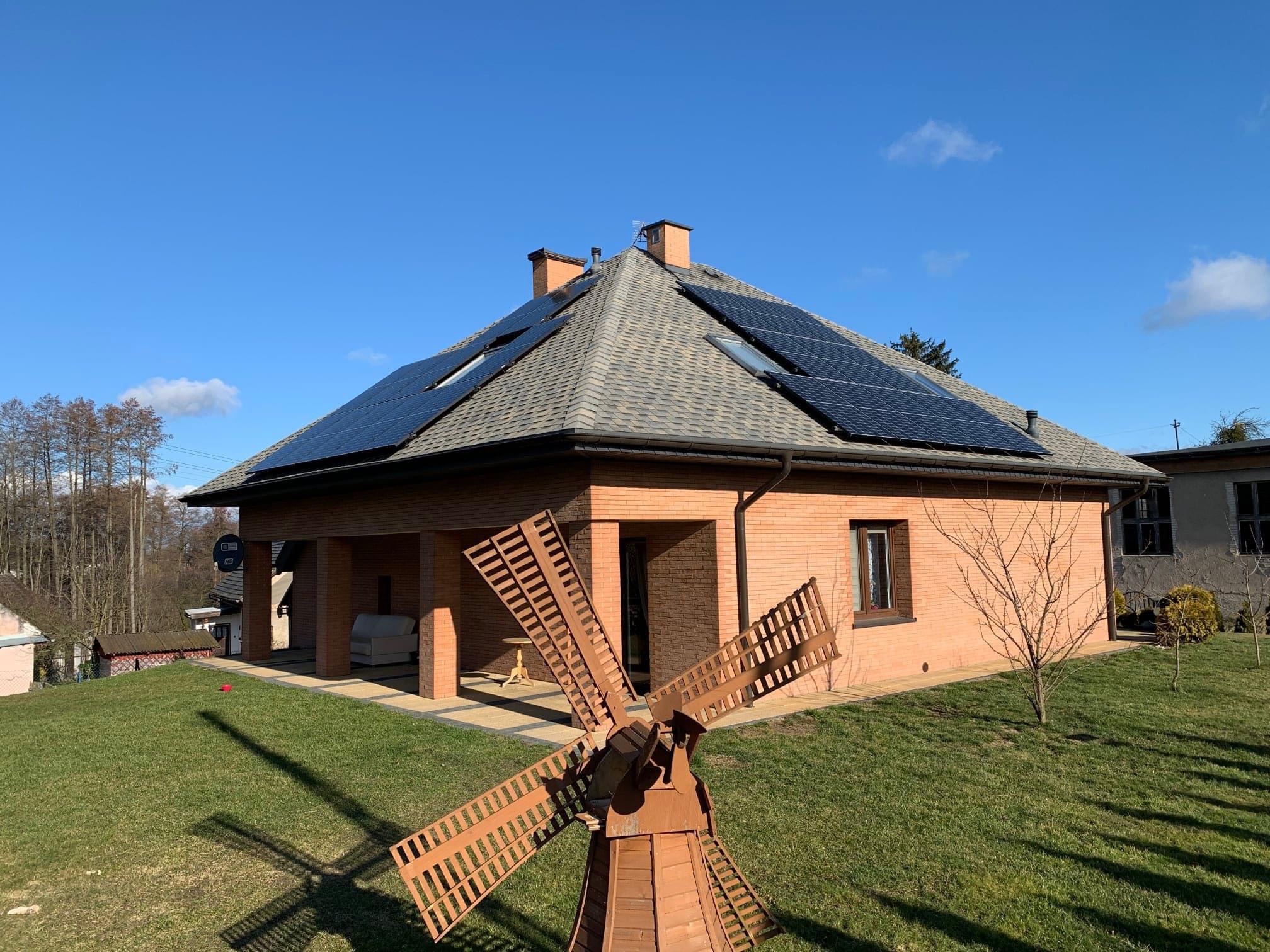 Dom z panelami słonecznymi na dachu, widoczny z perspektywy ogrodu z dekoracyjnym wiatrakiem na pierwszym planie, błękitne niebo w tle.