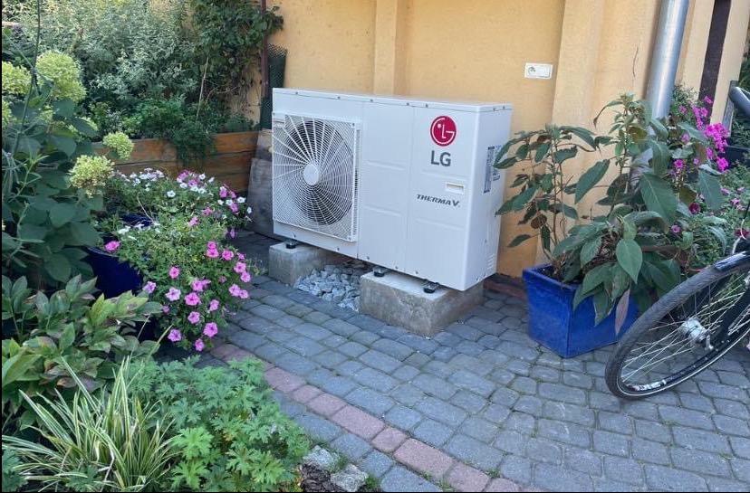 Biała pompa ciepła LG Therma V, ustawiona na betonowych blokach w ogrodzie, otoczona kwiatami i roślinami w donicach. Widoczny fragment roweru.