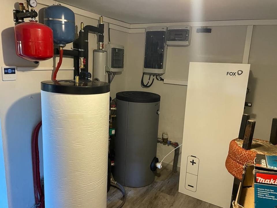 W kotłowni biały zbiornik z czerwonym i niebieskim naczyniem wzbiorczym, szary zbiornik oraz biały panel sterowania Fox. Widoczna instalacja rurowa i skrzynki elektryczne.