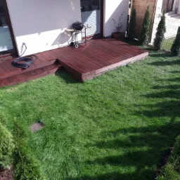 Świeżo założony trawnik z rolki otoczony tujami, z drewnianym tarasem i grillem przy białym budynku.