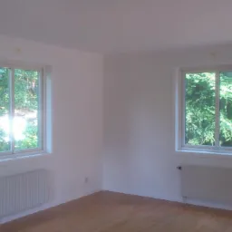 Jasne, puste pomieszczenie po remoncie z dwoma oknami z widokiem na zieleń i białymi grzejnikami pod nimi.