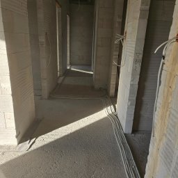 Hubert Żak - Wnętrze surowego budynku w trakcie instalacji elektrycznej: wiązka przewodów w peszlach ułożona wzdłuż ściany z betonu komórkowego, widoczne otwory drzwiowe i fragmenty instalacji w ścianach.