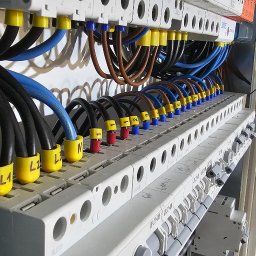 Hubert Żak - Szczegółowe ujęcie tablicy rozdzielczej z podłączonymi przewodami oznaczonymi kolorowymi końcówkami i opisami L1, L2, L3, N; widoczne białe kable z boku, uporządkowane za pomocą opaski zaciskowej.