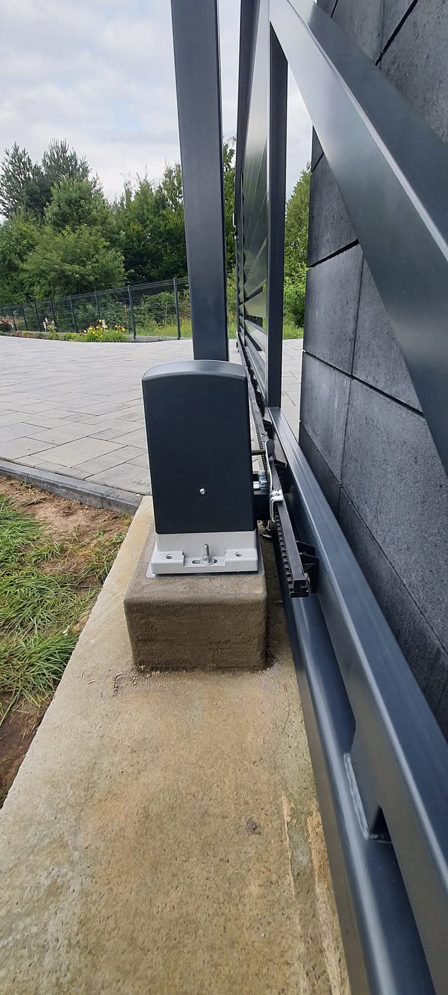 Szara, przesuwna brama wjazdowa z widocznym mechanizmem napędowym i łańcuchem, zamontowana na betonowej podstawie, z kostką brukową i zielenią w tle.