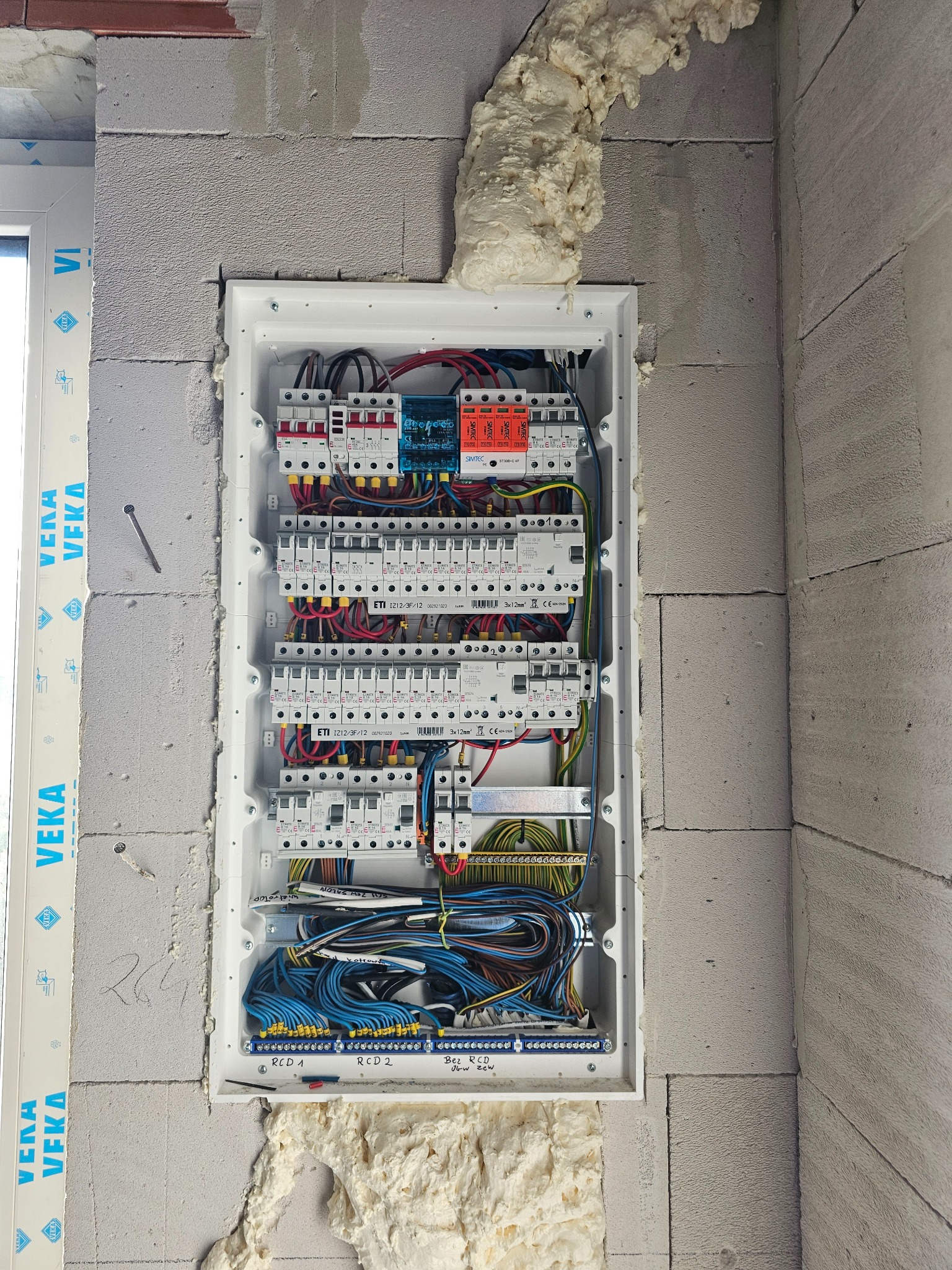 Rozdzielnica elektryczna z zabezpieczeniami, zamontowana w surowej ścianie z betonu komórkowego, uszczelniona pianką montażową, widoczne kable i bezpieczniki ETI.