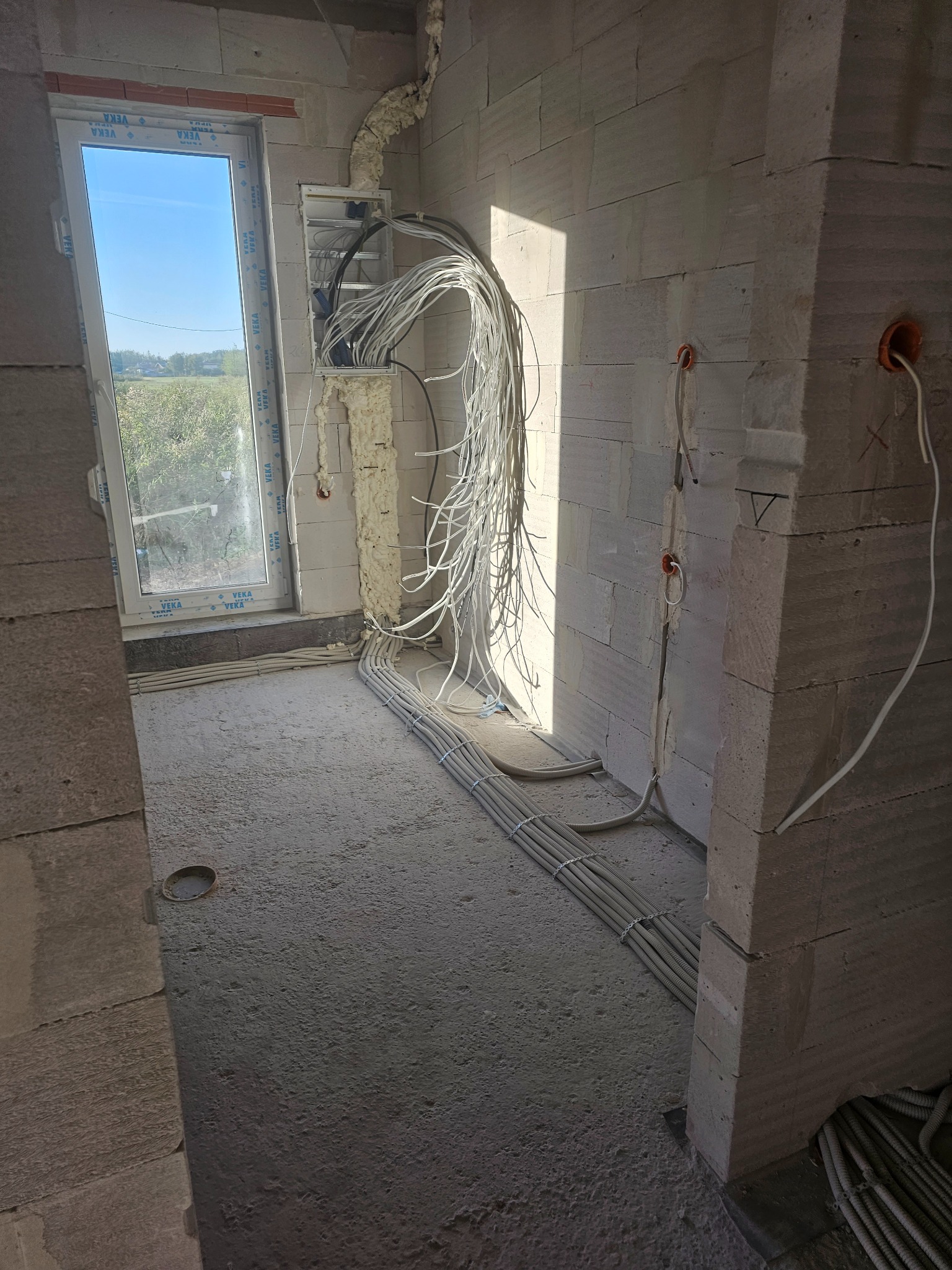 Surowy stan budynku w trakcie instalacji elektrycznej: kable w peszlach ułożone wzdłuż ściany i pod oknem, wiszące przewody z puszki elektrycznej zabezpieczone pianką montażową. Widoczne otwory...
