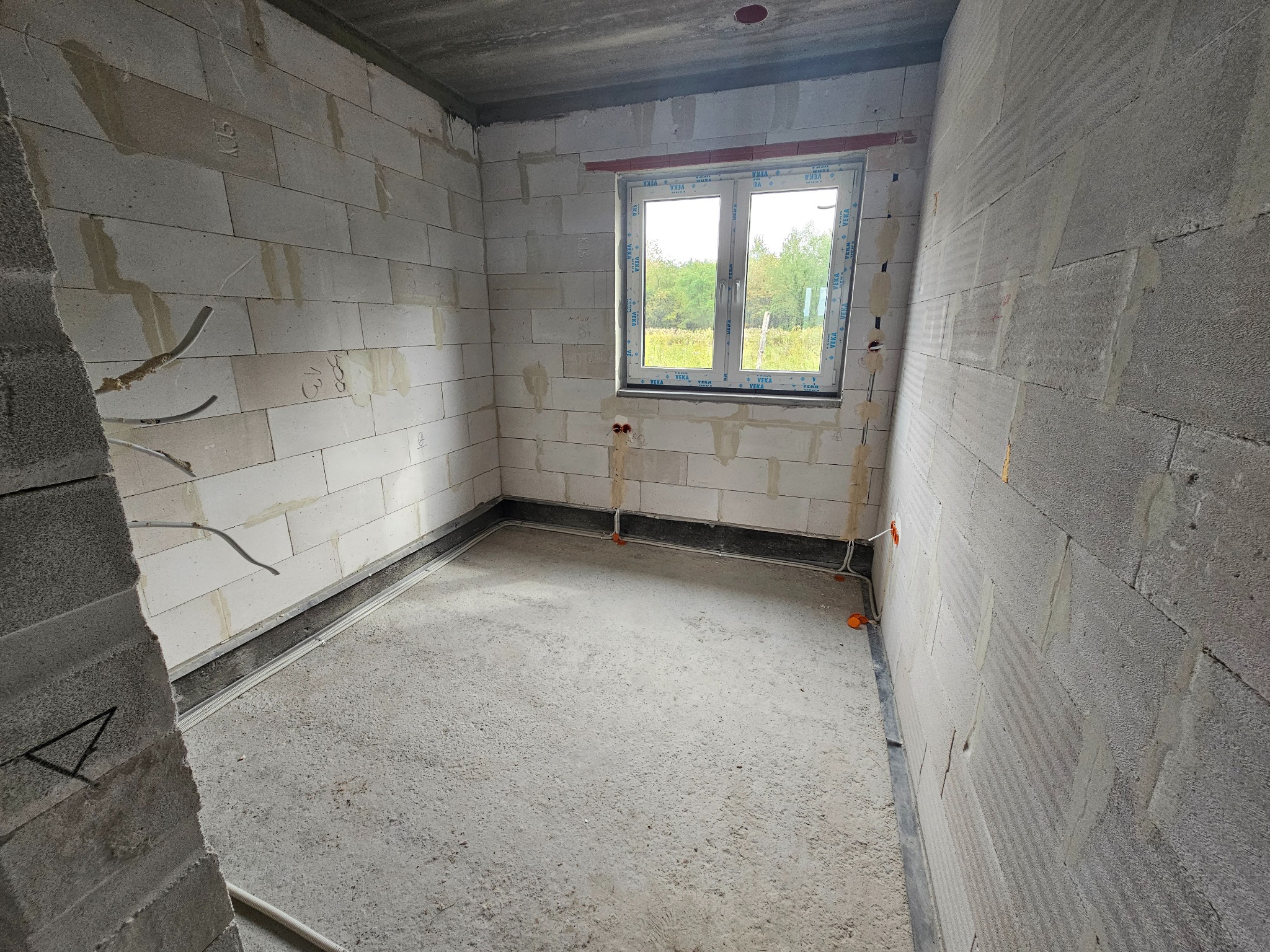 Surowe wnętrze w trakcie prac instalacyjnych: widoczne przewody elektryczne w ścianach z betonu komórkowego, okno z widokiem na zieleń, surowy beton na podłodze i suficie.